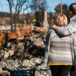 A couple standing together, embracing, as they survey the aftermath of a natural disaster with burned vehicles and debris.