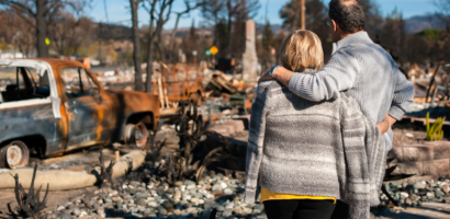 A couple standing together, embracing, as they survey the aftermath of a natural disaster with burned vehicles and debris.