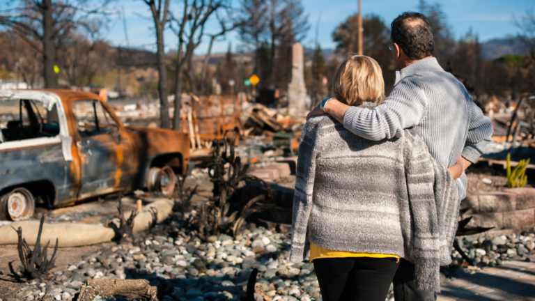 A couple standing together, embracing, as they survey the aftermath of a natural disaster with burned vehicles and debris.