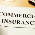 A hand holding a document with the words "Commercial Insurance" printed on it, accompanied by a pen and a keyboard on a desk.