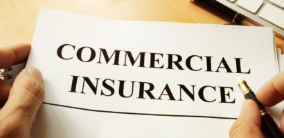 A hand holding a document with the words "Commercial Insurance" printed on it, accompanied by a pen and a keyboard on a desk.